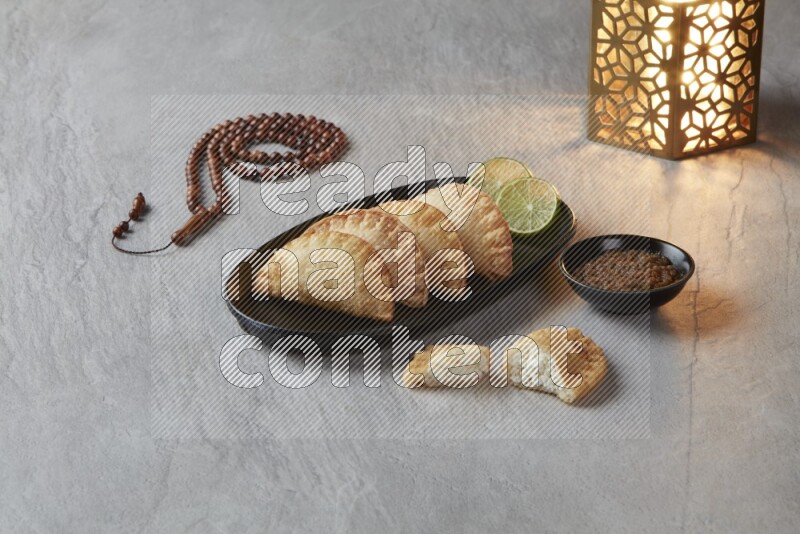 Four fried sambosas in an oval shaped black plate, beside a cut cheese sambosa, a brown misbaha and a golden lantern on a gray background