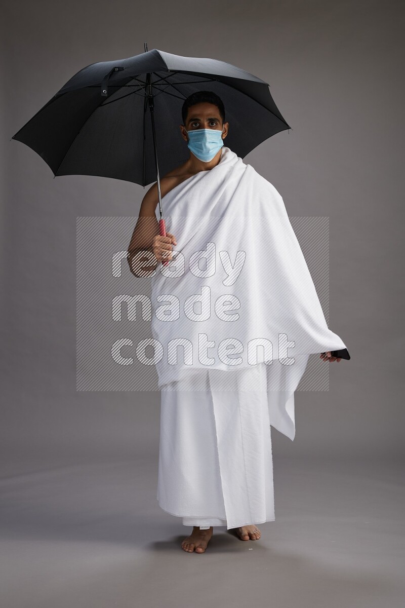 A man wearing Ehram and mask Standing holding umbrella on gray background on gray background