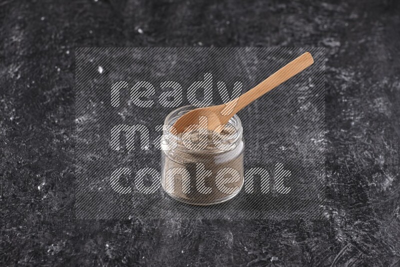 A glass jar full of black pepper powder and a wooden spoon on a textured black flooring
