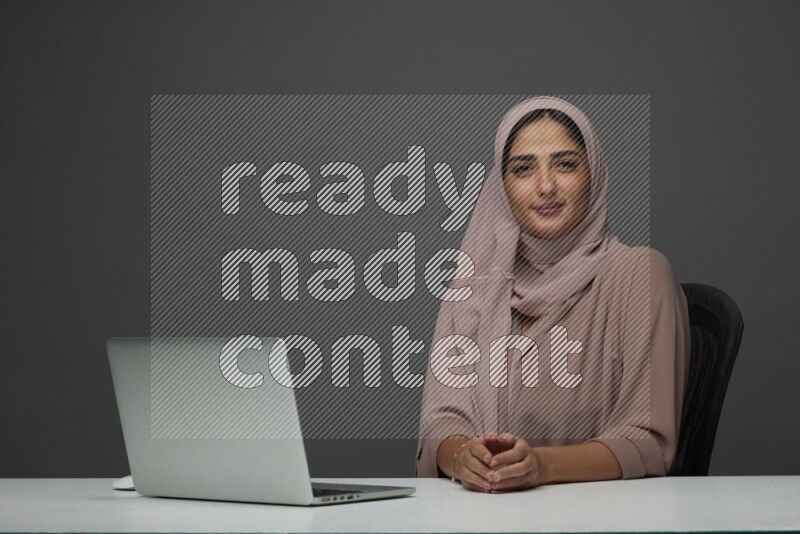 A Saudi woman Setting on her desk on a Gray Background wearing Brown Abaya with Hijab