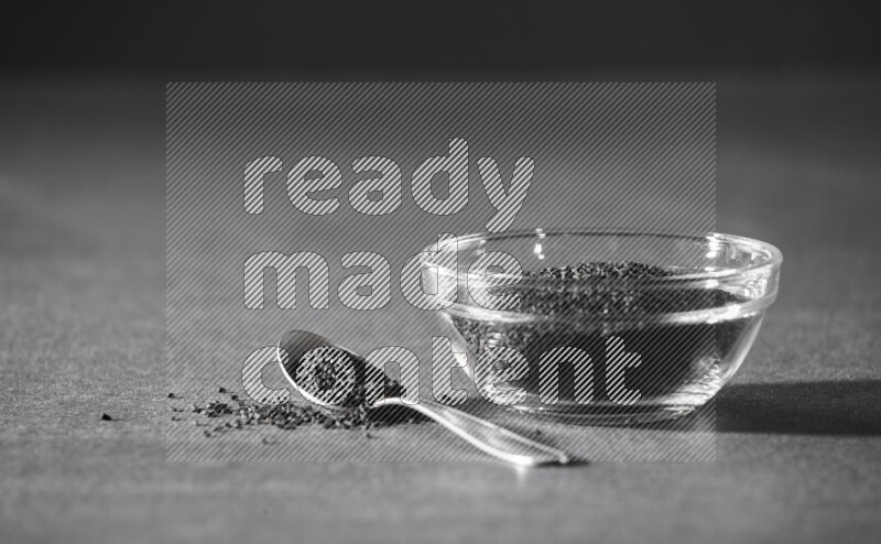 وعاء زجاجي ممتلئ ببذور حبة البركة مع ملعقة معدنية مليئة بحبة البركة علي خلفية سوداء