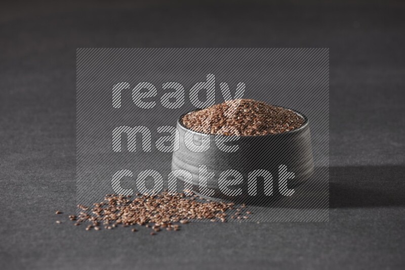 A black pottery bowl full of flaxseeds surrounded by the seeds on a black flooring