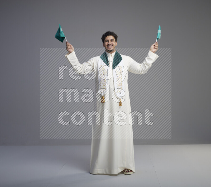 A Saudi man standing wearing thob and saudi flag scarf and holding small saudi flag on gray background