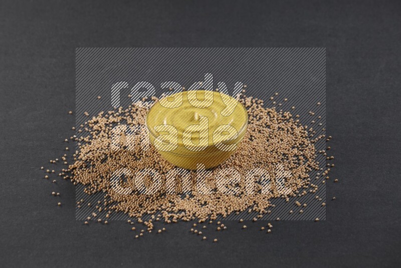 A glass bowl full of mustard paste with mustard seeds underneath on black flooring