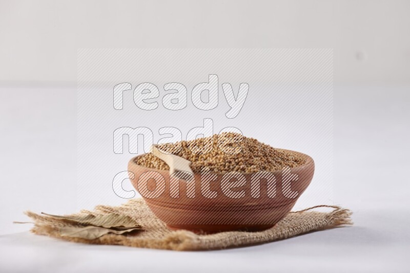 A wooden bowl full of mustard seeds with a wooden spoon on the seed set on burlap fabric on a white flooring