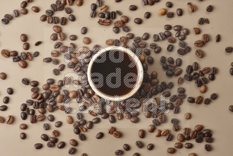 A beige pottery cup of coffee surrounded by roasted coffee beans on beige background
