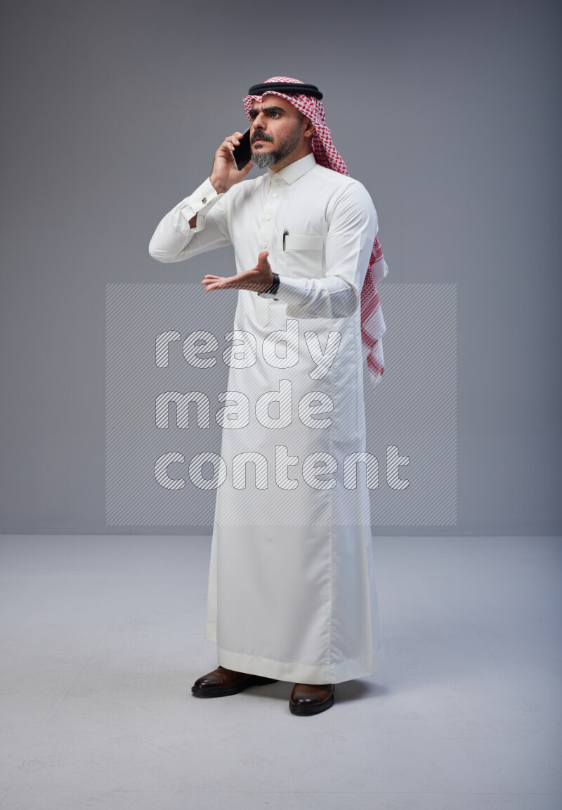 Saudi man Wearing Thob and red Shomag standing talking on phone on Gray background
