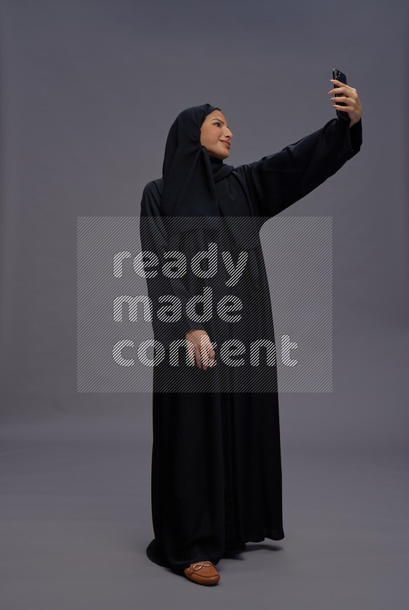Saudi woman wearing Abaya standing taking selfie on gray background