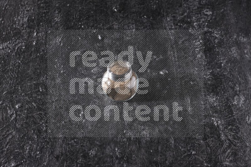 A glass spice jar full of black pepper powder on textured black flooring
