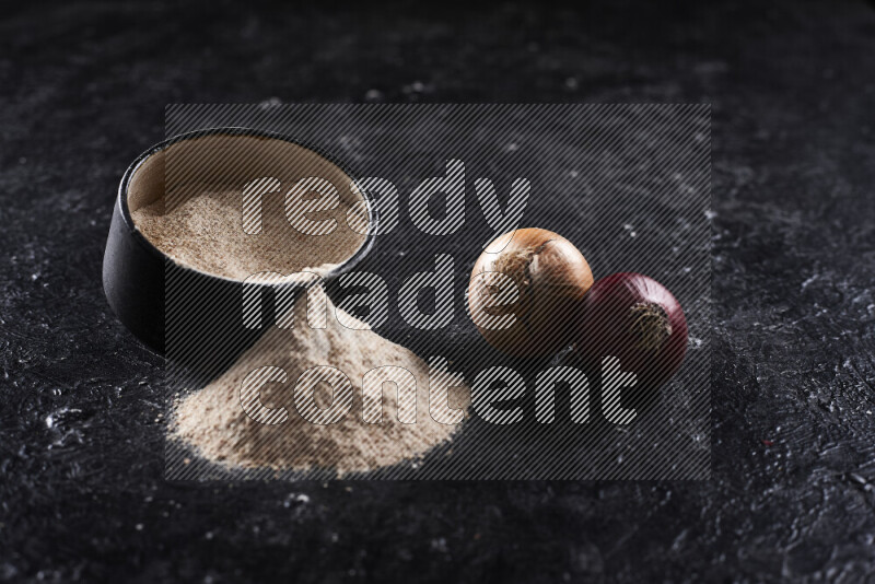 A black pottery bowl full of onion powder with fallen powder from it on black background