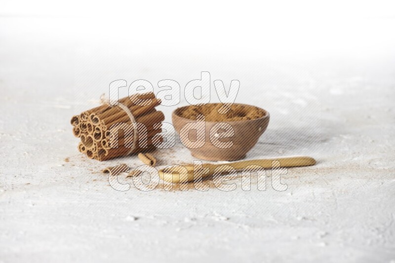 Cinnamon sticks stacked and bounded beside a wooden bowl full of cinnamon powder and a wooden spoon full of powder on white background