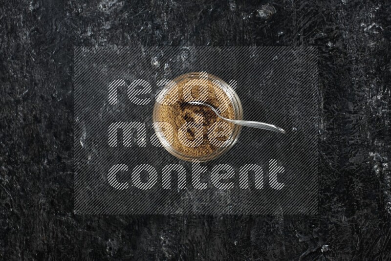 A glass jar and a metal spoon full of cumin powder on a textured black flooring