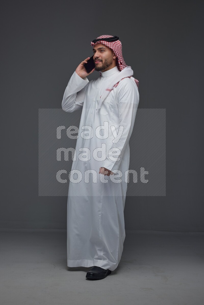 A Saudi man wearing Thobe and Shmagh talking in the phone eye level on a gray background