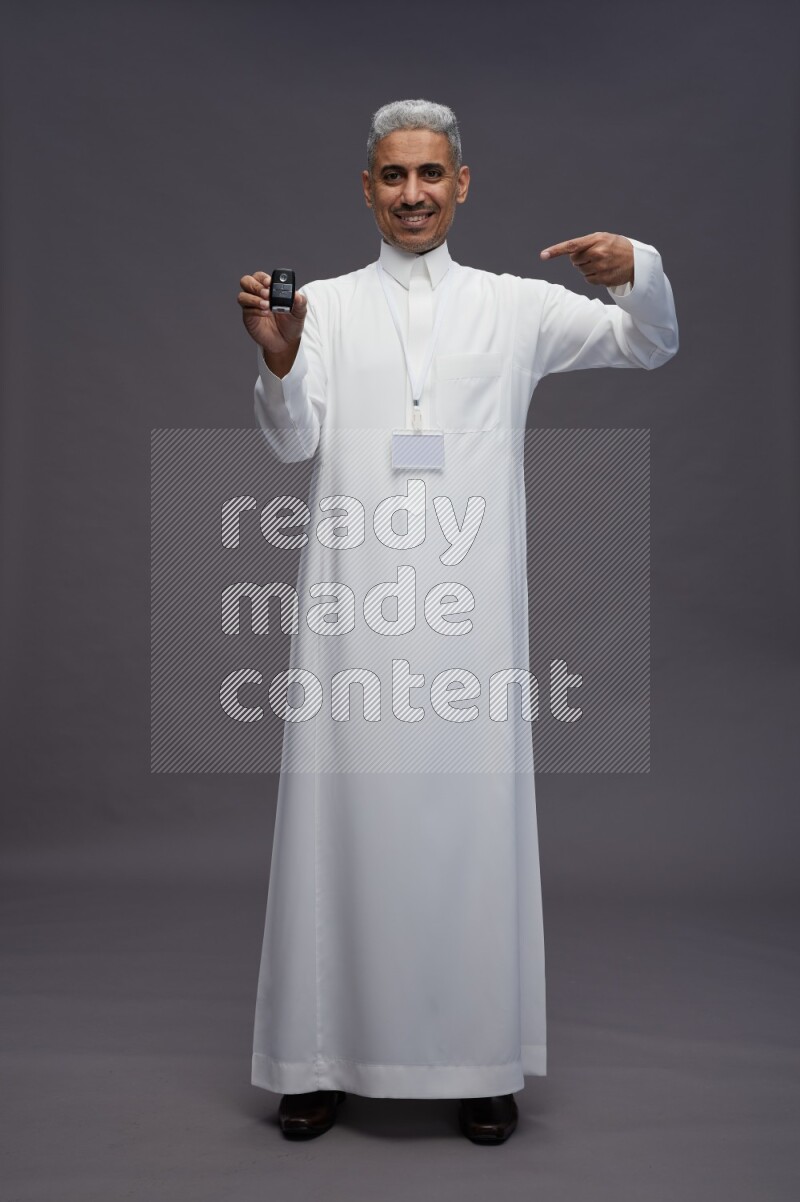 Saudi man wearing thob with neck strap employee badge standing holding key car on gray background
