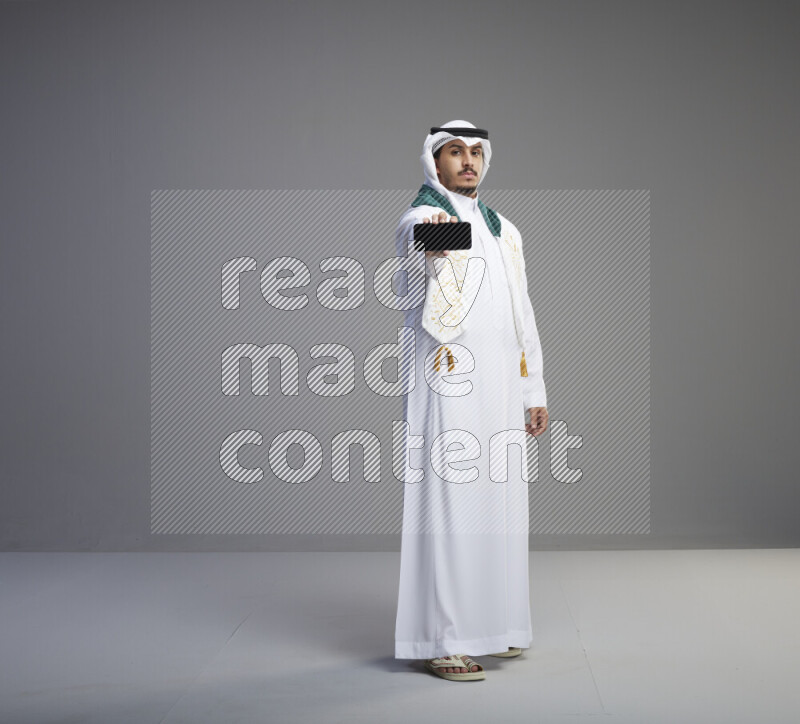 A Saudi man standing wearing thob and white shomag with flag scarf on his neak showing phone on gray background