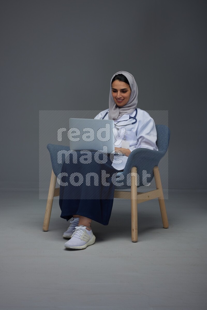 A doctor wearing a light gray head scarf sitting on a dark grey chair and using a laptop eye level on a grey background