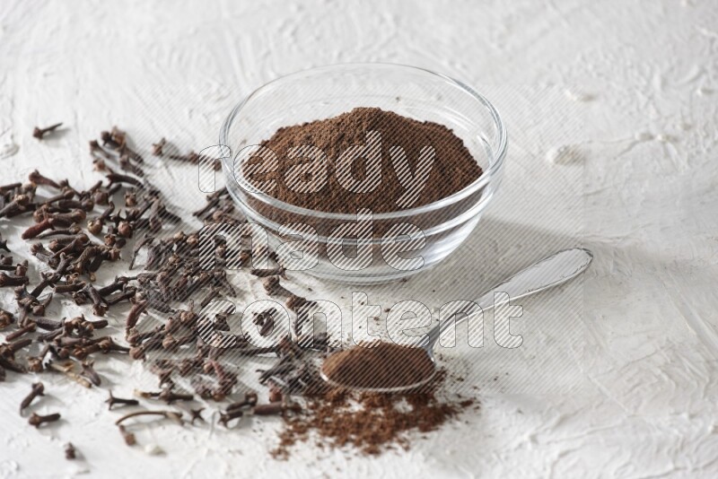 A glass bowl and a metal spoon full of cloves powder with cloves grains spread on a textured white flooring