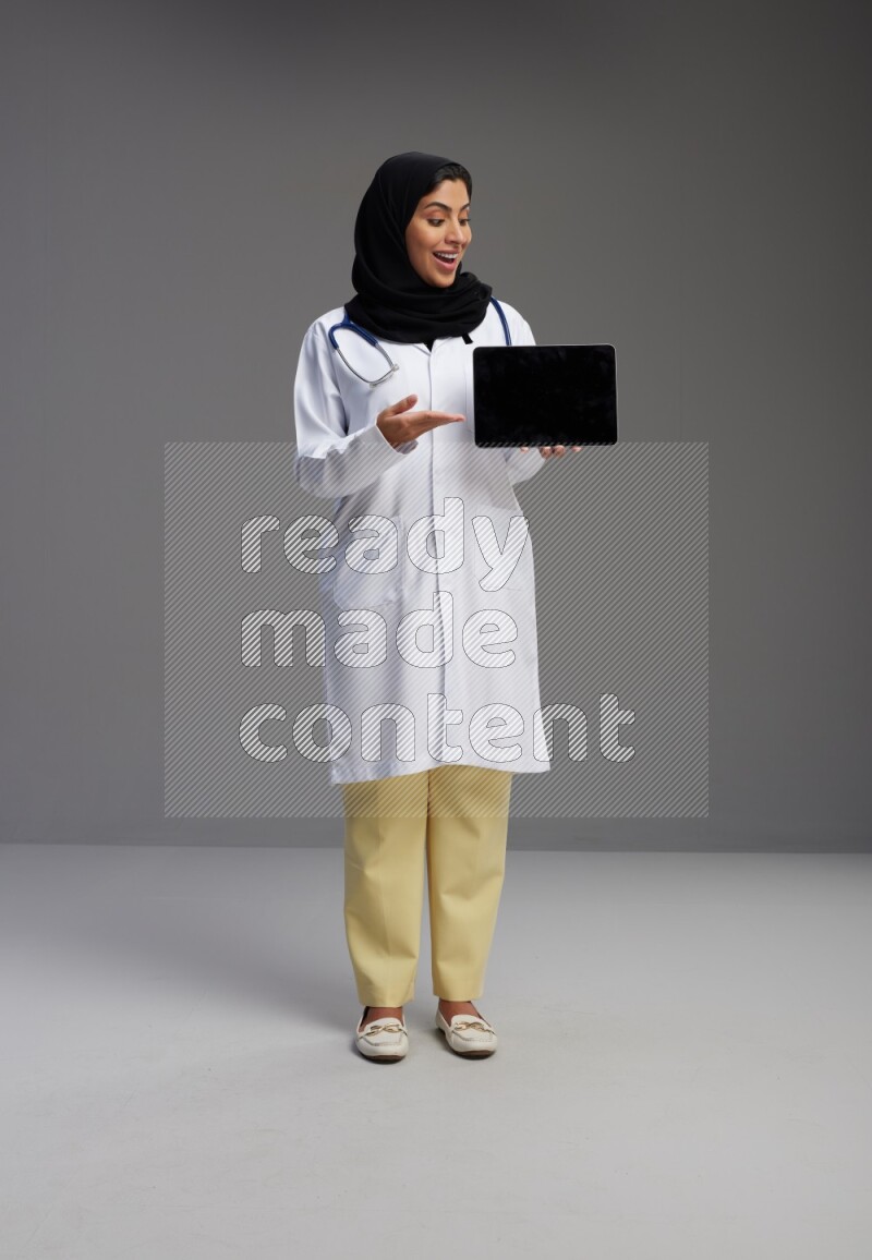 Saudi woman wearing lab coat with stethoscope standing showing tablet to camera with sign in the back on Gray background