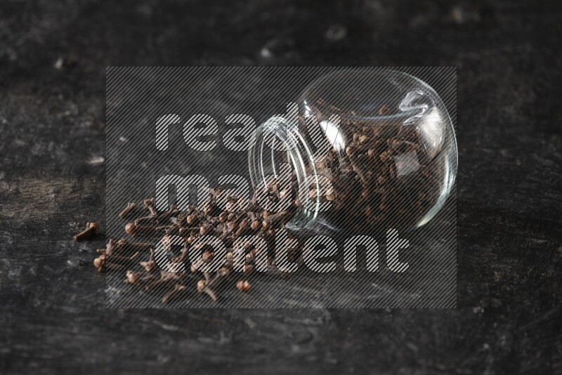 A flipped glass spice jar full of cloves and cloves came out of it on textured black flooring