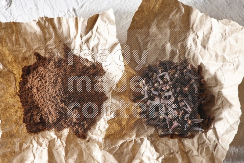 Cloves powder and cloves on 2 crumpled pieces of paper on a textured white flooring
