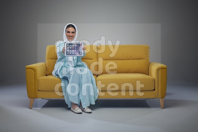 A Saudi woman wearing a light blue Abaya and white head scarf sitting on a yellow sofa and showing her tablet's screen while pointing to it eye level on a grey background