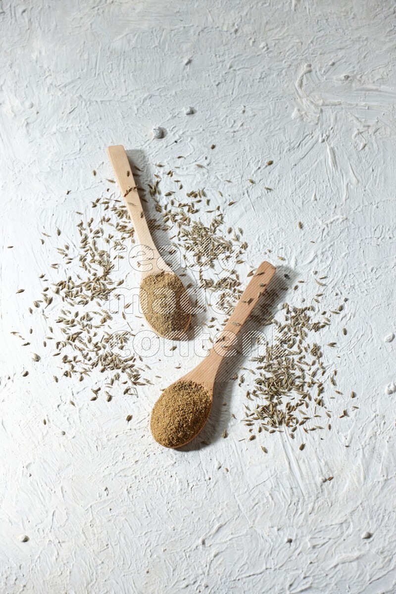 2 Wooden spoons full of cumin powder and cumin seeds on textured white flooring