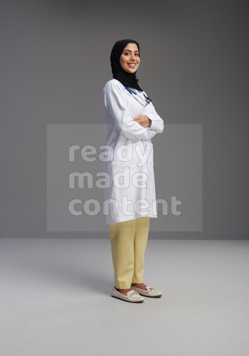 Saudi woman wearing lab coat with stethoscope standing with crossed arms on Gray background