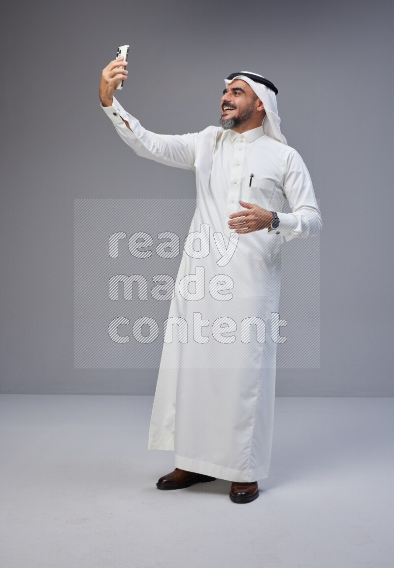 Saudi man Wearing Thob and white Shomag standing taking selfie on Gray background