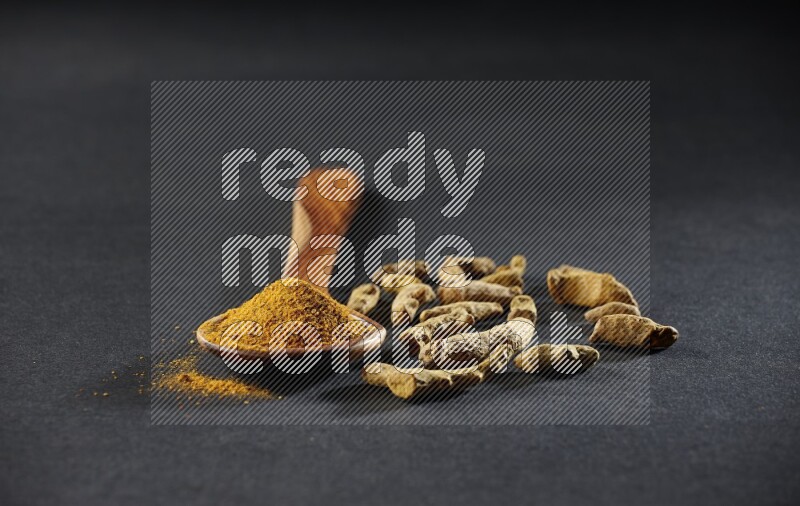 A wooden ladle full of turmeric powder with dried turmeric fingers on black flooring
