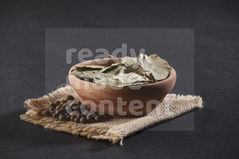 A wooden bowl filled with dried bay leaves on a piece of burlap with bunch of allspice berries on black flooring
