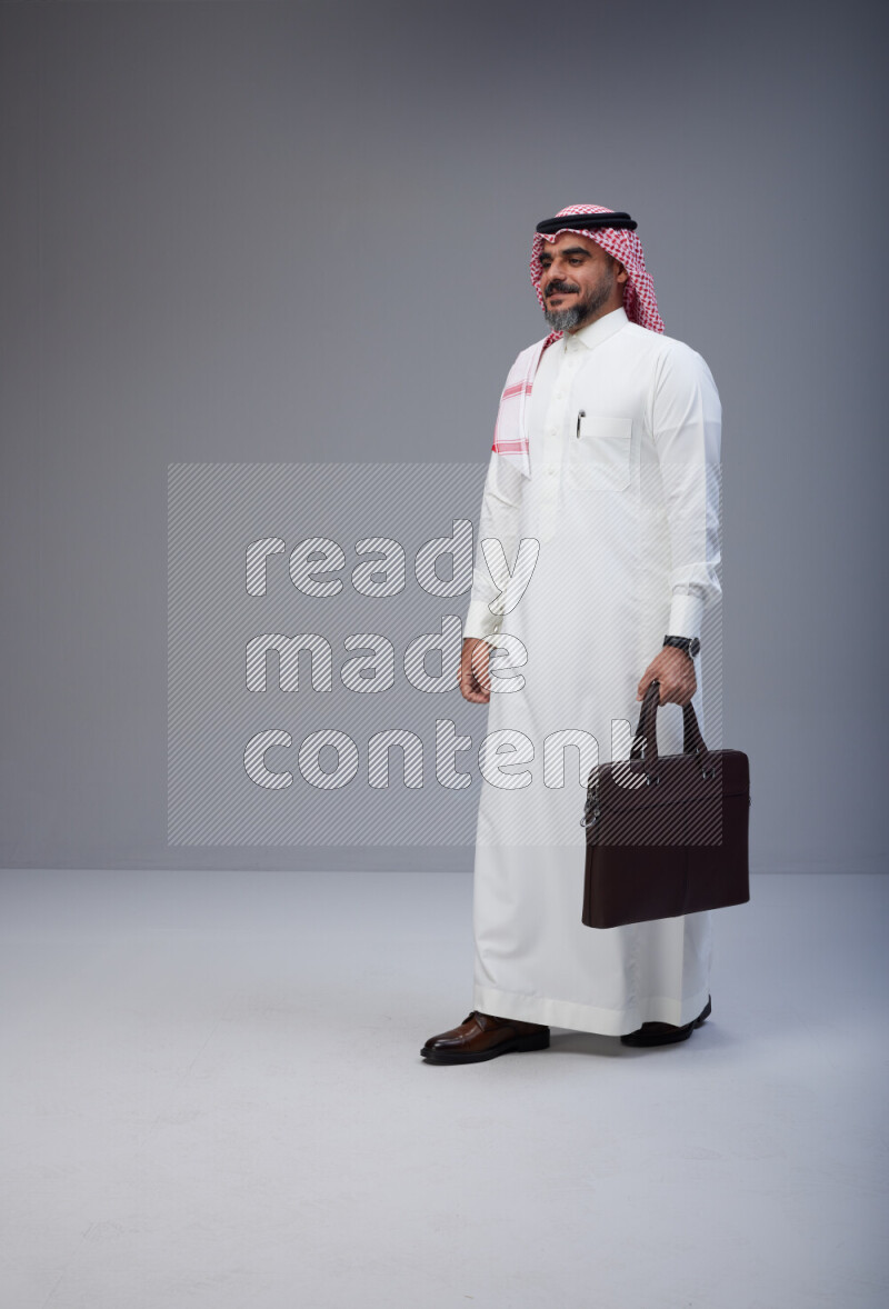 Saudi man Wearing Thob and red Shomag standing holding bag on Gray background