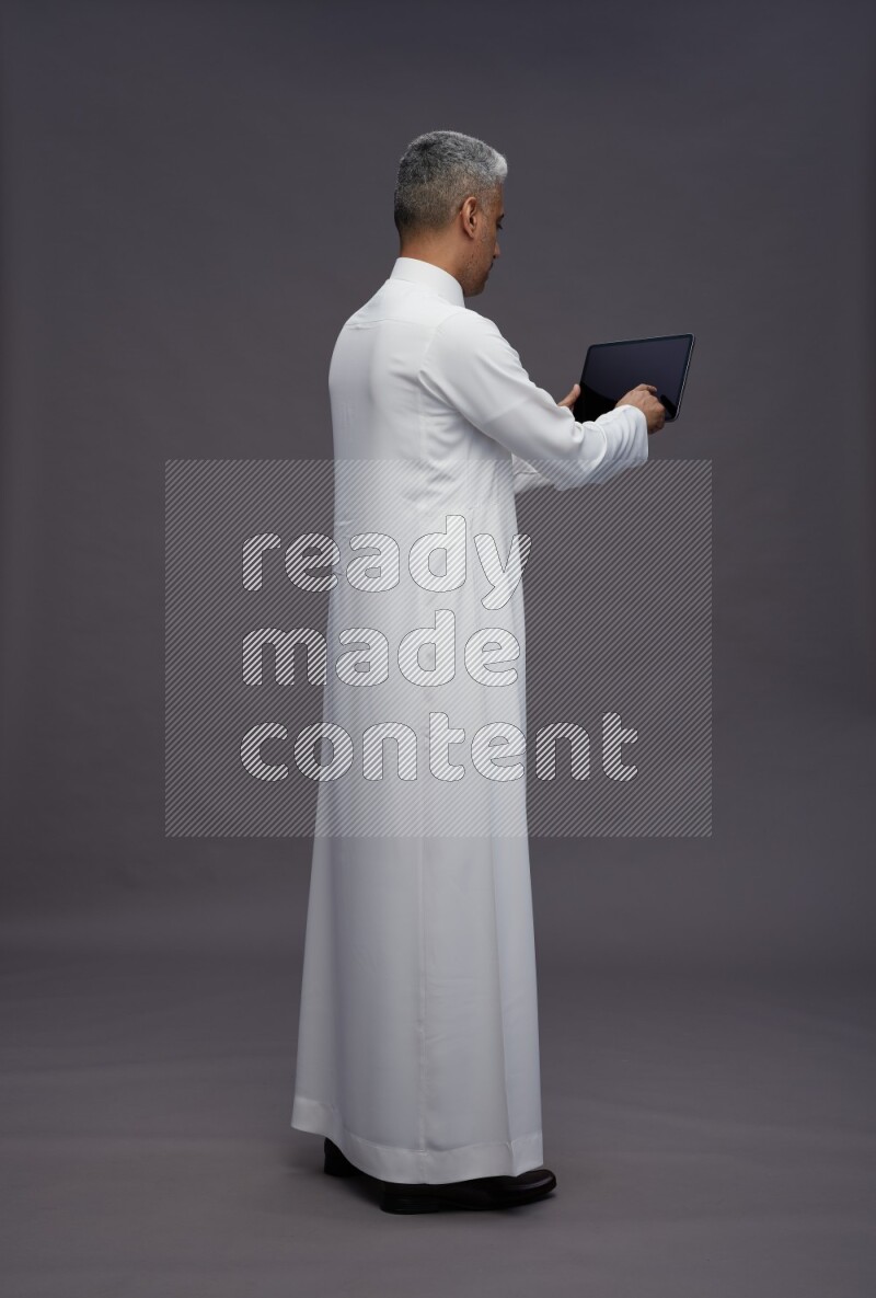 Saudi man wearing thob standing working on tablet on gray background