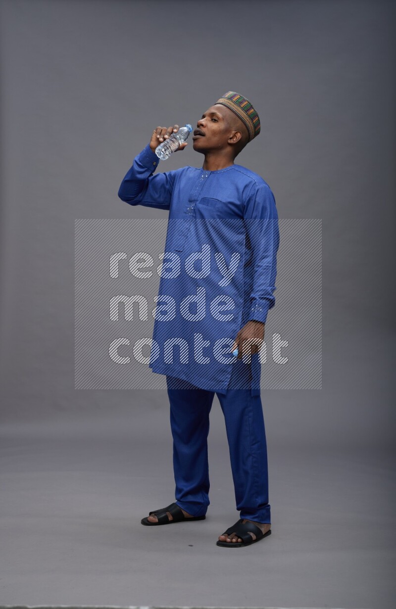 Man wearing Nigerian outfit standing drinking water on gray background