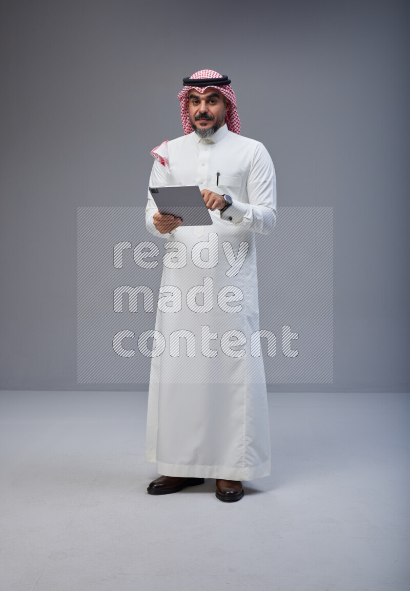 Saudi man Wearing Thob and red Shomag standing working on tablet on Gray background