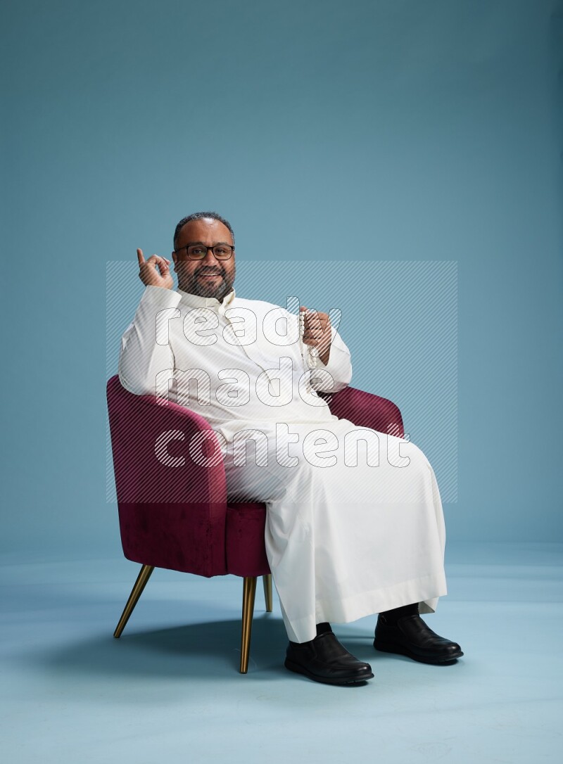 Saudi Man without shimag sitting on chair Interacting with the camera on blue background