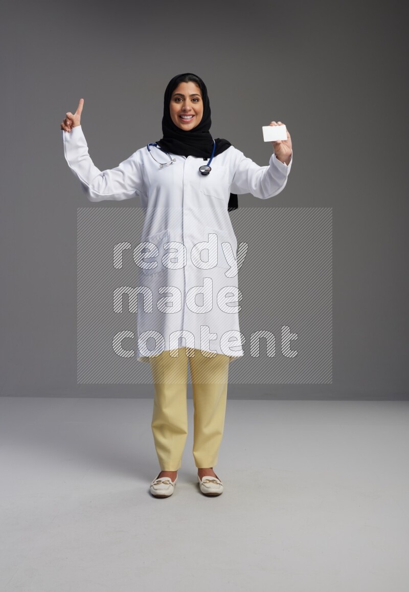 Saudi woman wearing lab coat with stethoscope standing holding ATM card on Gray background