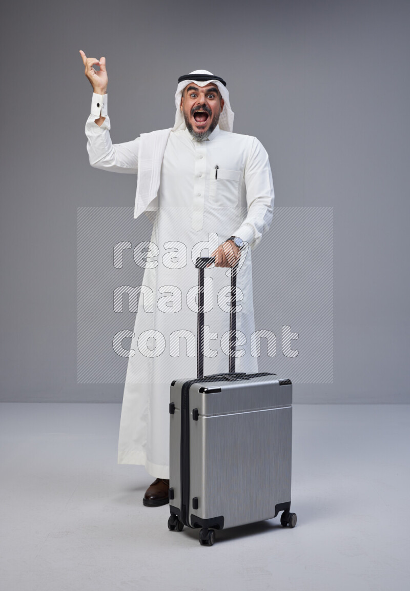 Saudi man wearing Thob and white Shomag standing holding Travel bag on Gray background