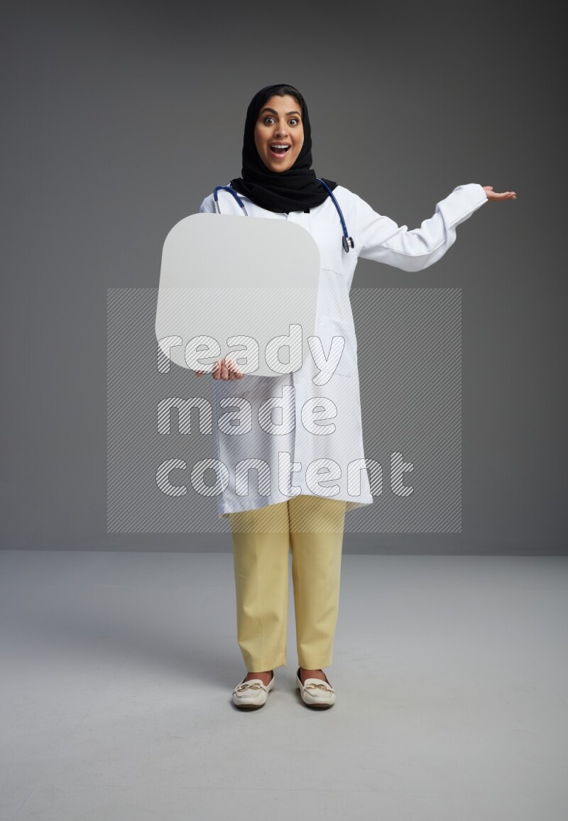 Saudi woman wearing lab coat with stethoscope standing holding social media sign on Gray background