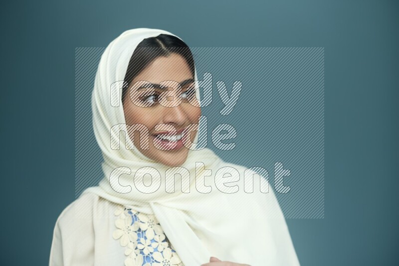 A Saudi woman posing in a blue background wearing an off-white Abaya with hijab