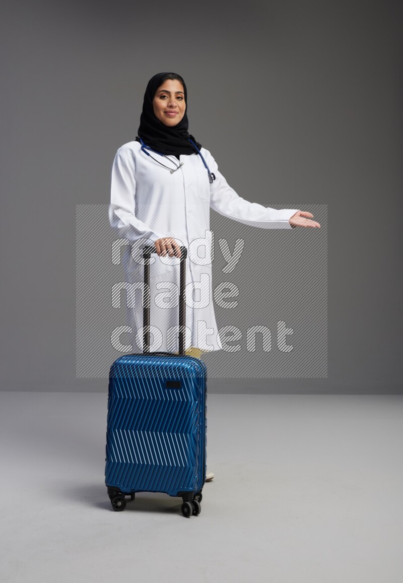 Saudi woman wearing lab coat with stethoscope standing holding Travel bag on Gray background