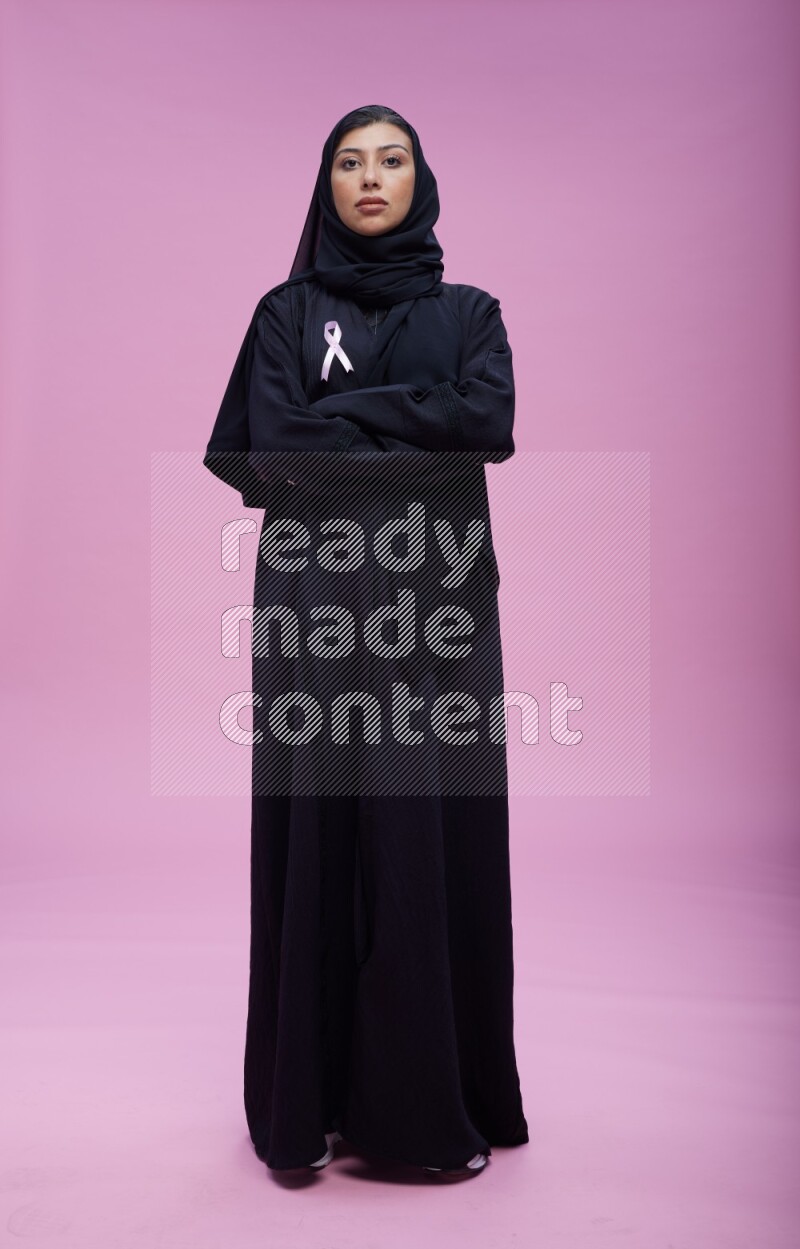 Saudi woman wearing pink ribbon on Abaya standing crossed arms on pink background