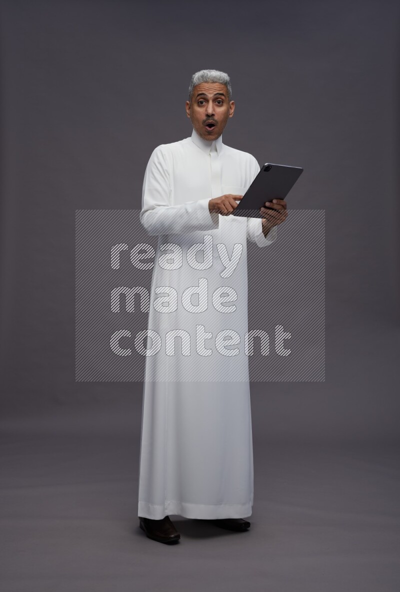 Saudi man wearing thob standing working on tablet on gray background