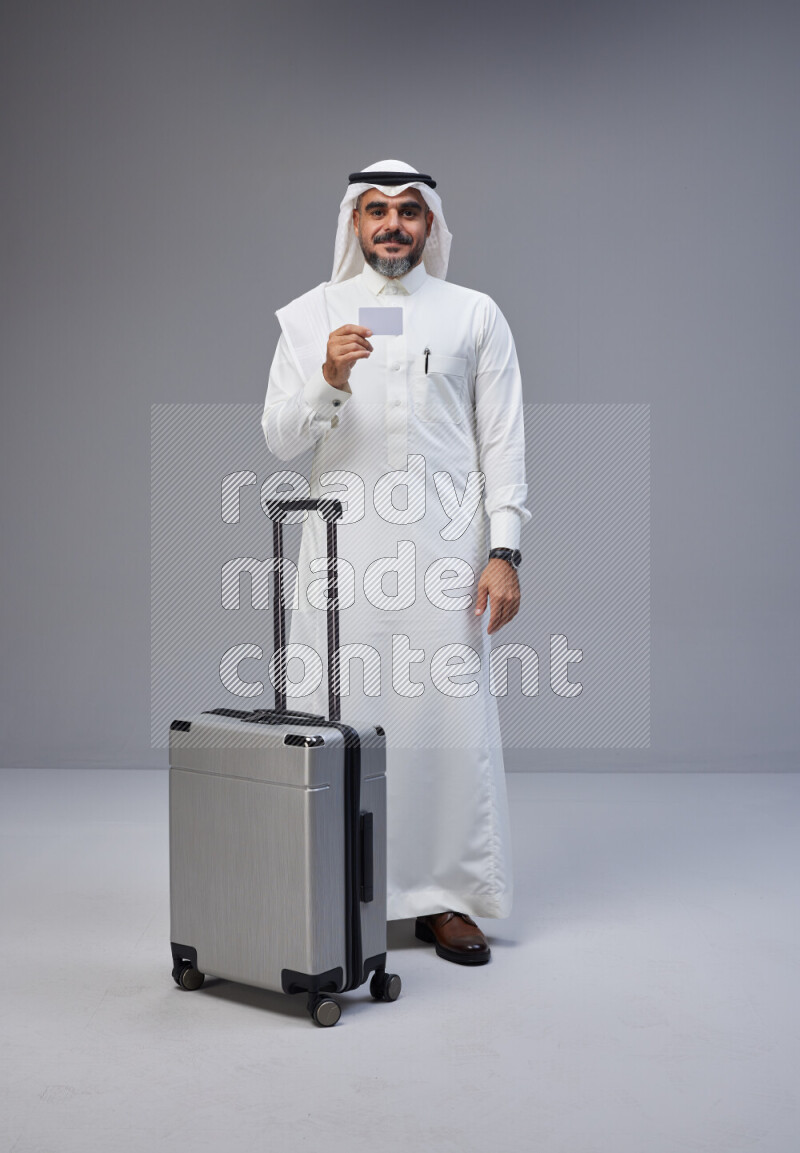 Saudi man wearing Thob and white Shomag standing holding Travel bag and ATM card on Gray background