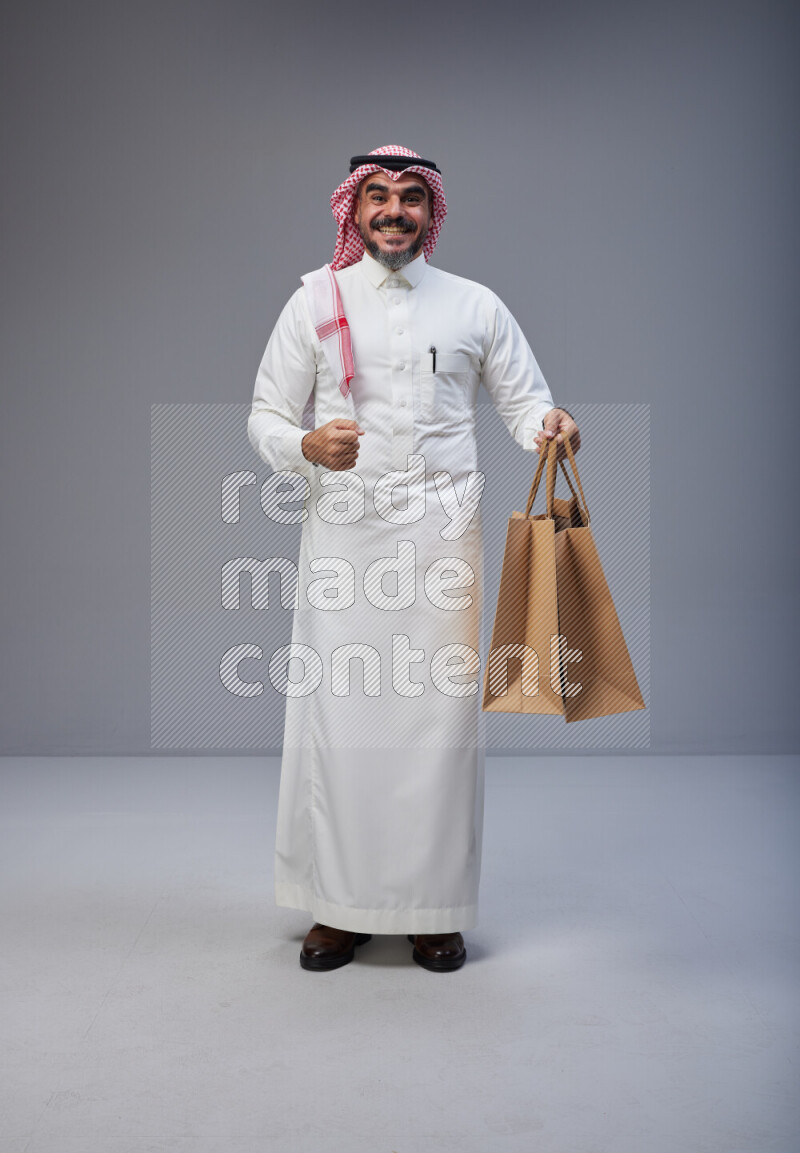 Saudi man Wearing Thob and red Shomag standing holding shopping bag on Gray background
