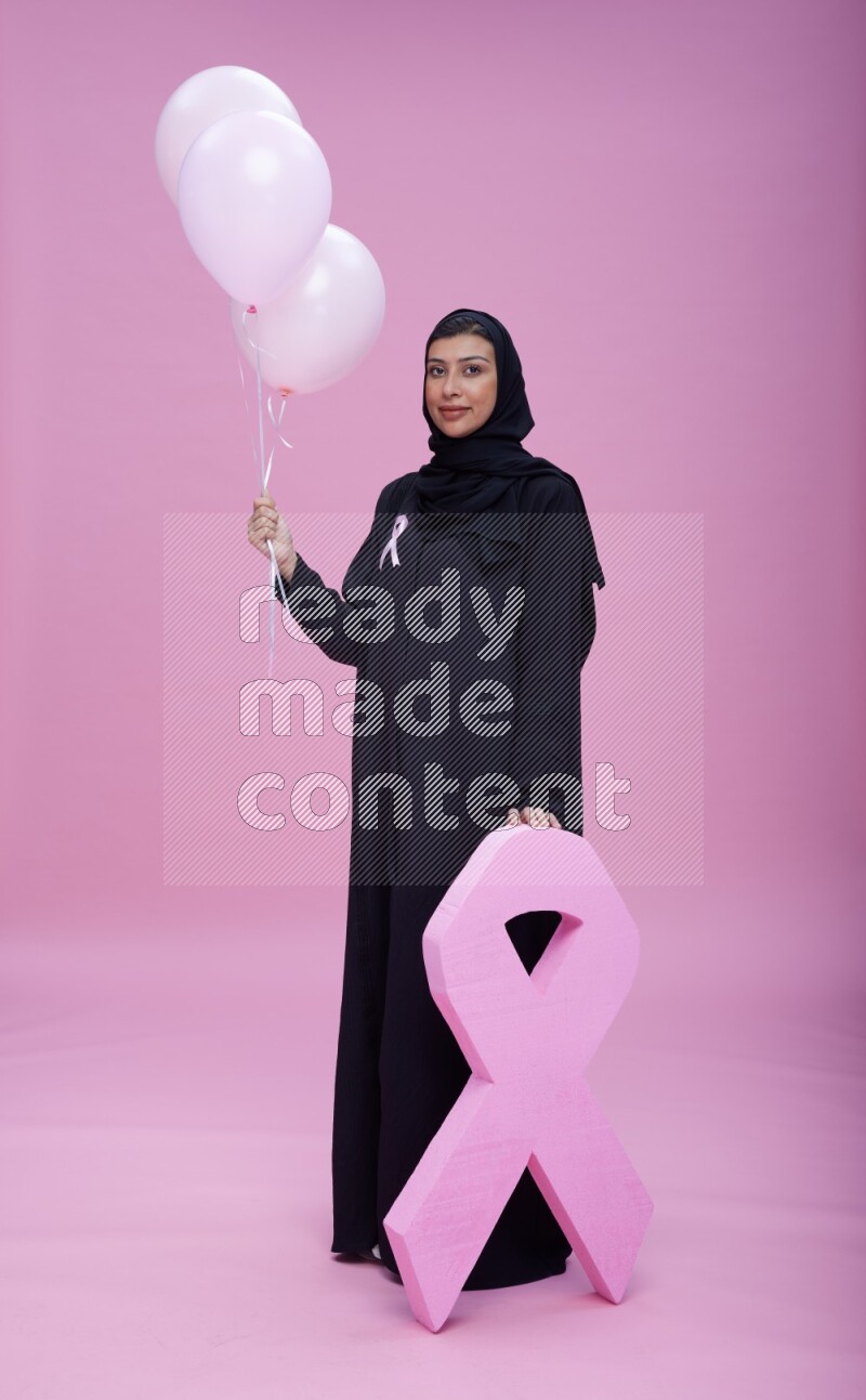 Saudi woman wearing pink ribbon on Abaya standing holding balloons and awareness sign on pink background