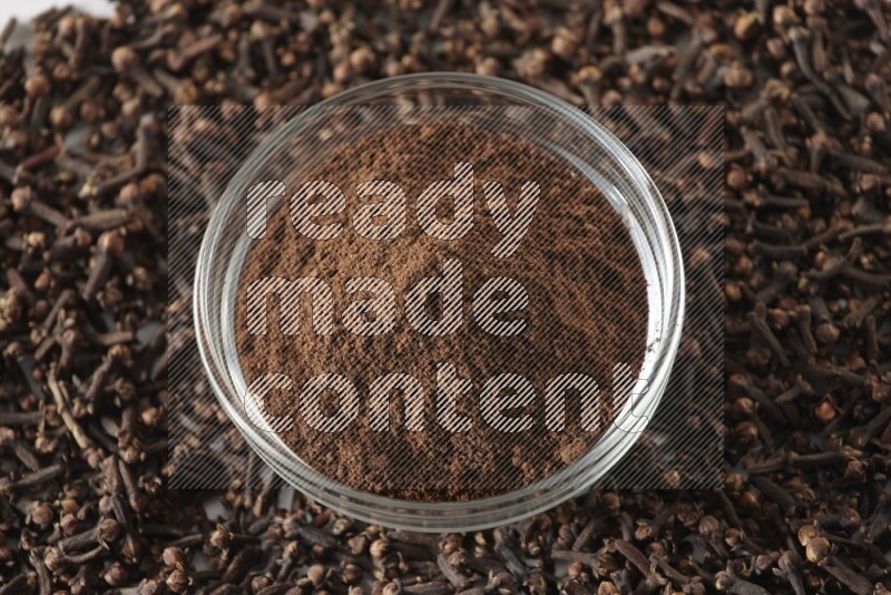 وعاء زجاجي ممتلئ ببودرة القرنفل وحبوب القرنفل متناثرة على أرضية بيضاء