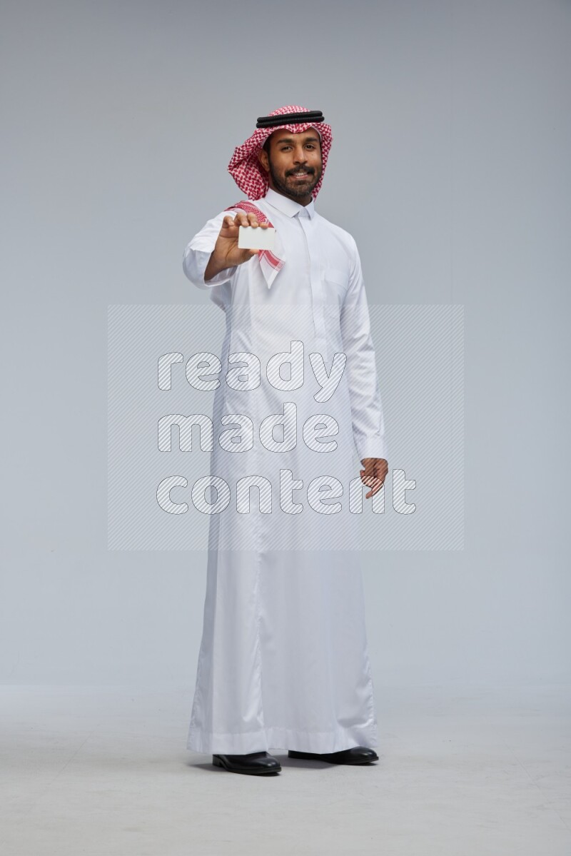 Saudi man wearing thob and shomag standing holding ATM card on gray background