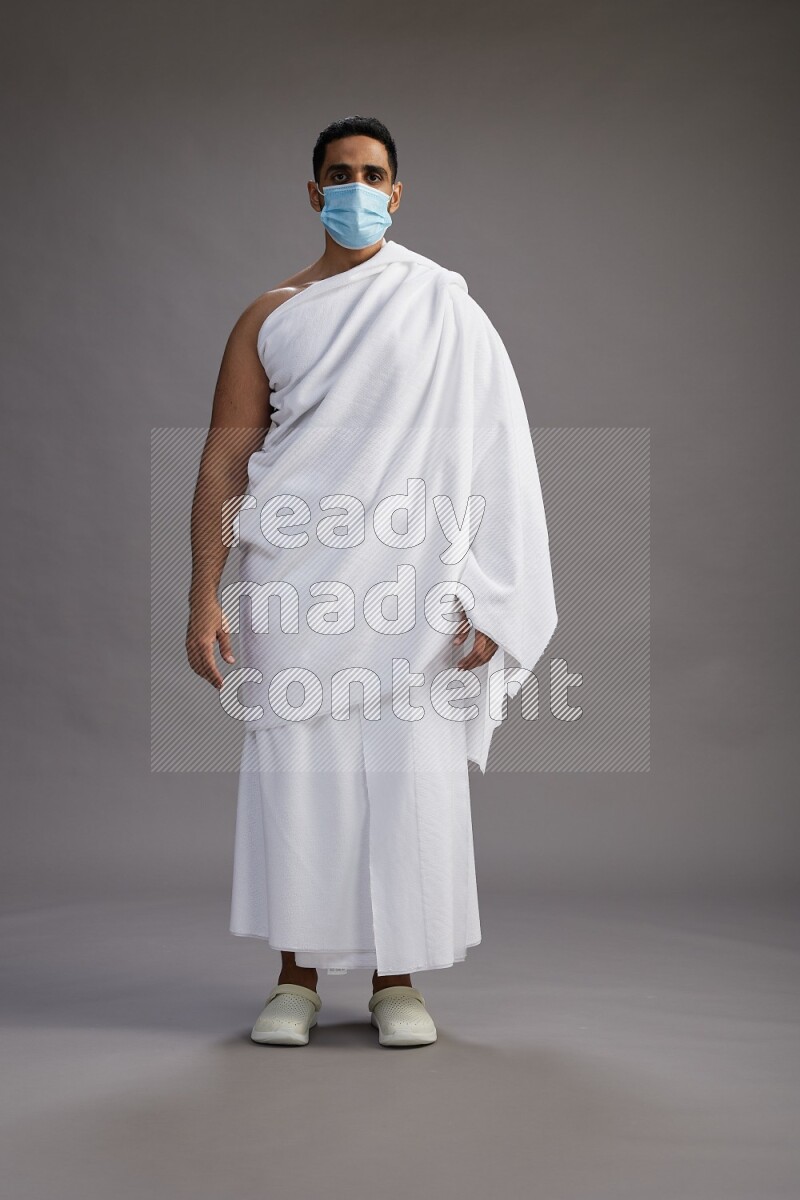 A man wearing Ehram with face mask Standing Interacting with the camera on gray background