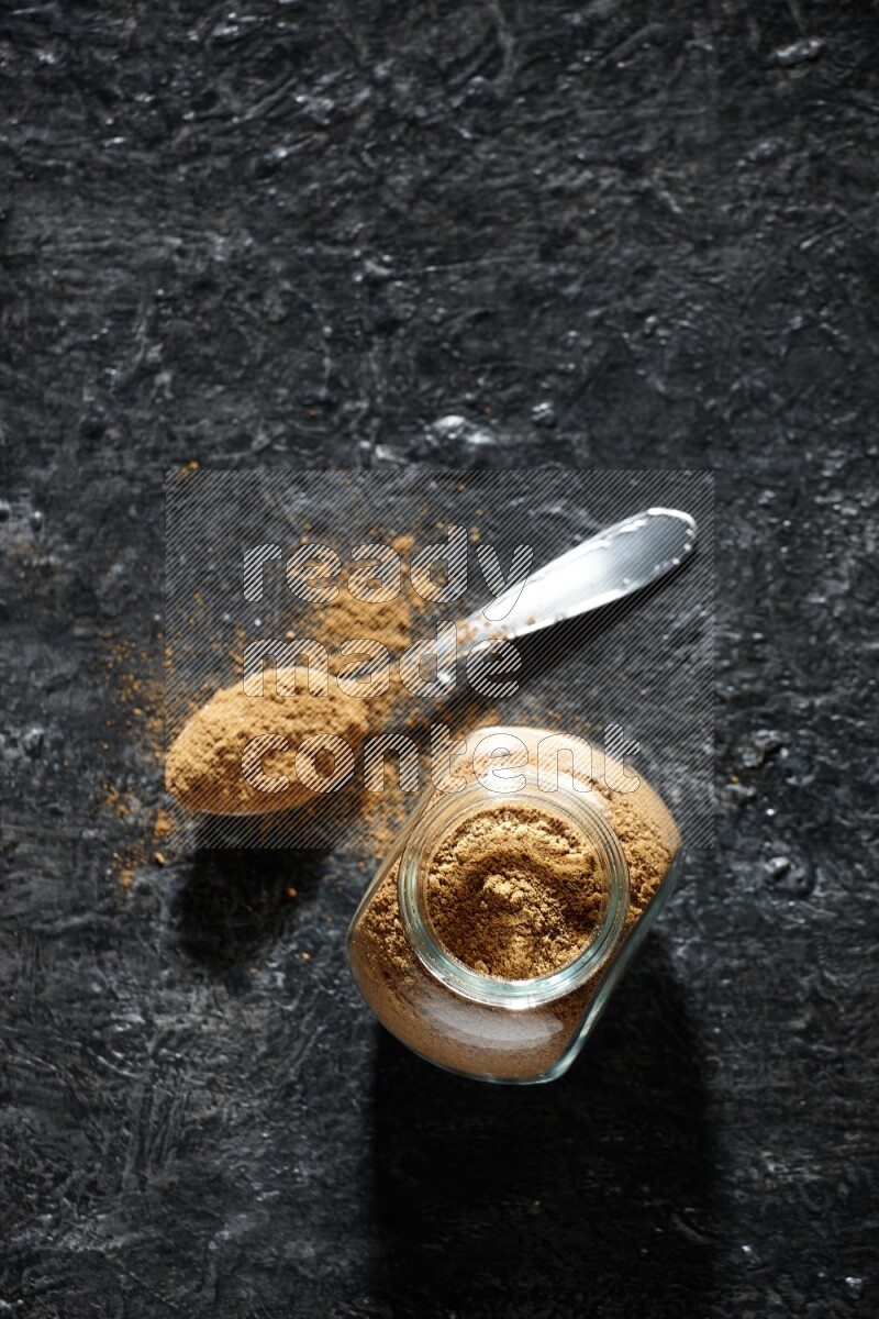 A glass spice jar and metal spoon full of allspice powder on a textured black flooring