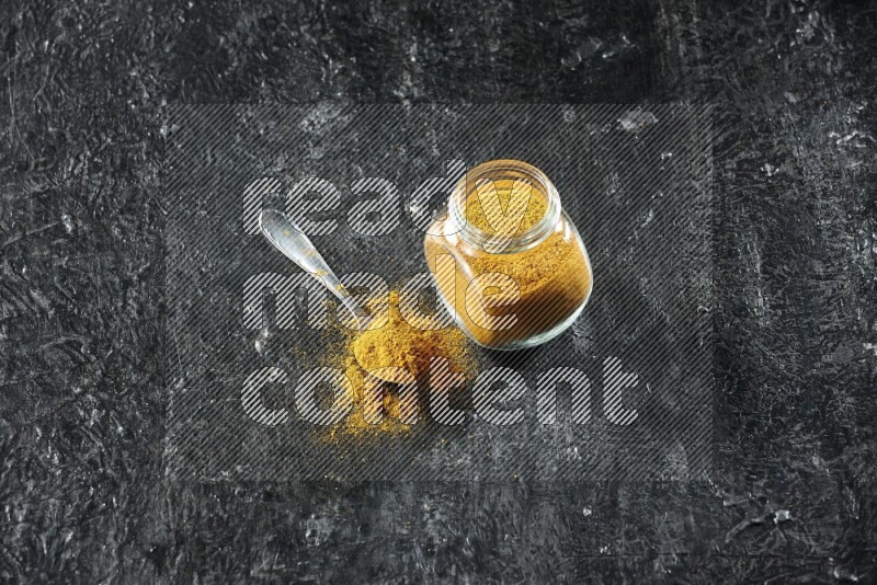 A glass spice jar and metal spoon full of turmeric powder on textured black flooring